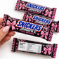 A hand holds a custom Snickers Amara's 10th Birthday chocolate bar wrapper, with three more identical wrappers arranged behind it on a white background. The wrapper features a pink leopard print, pink bows, and the blue and white Snickers logo.
