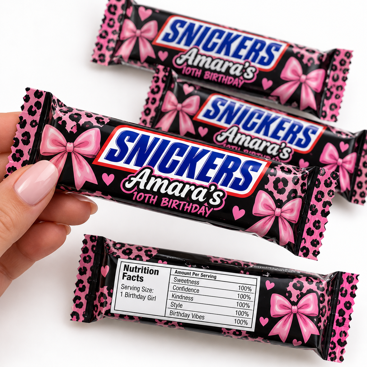 A hand holds a custom Snickers Amara's 10th Birthday chocolate bar wrapper, with three more identical wrappers arranged behind it on a white background. The wrapper features a pink leopard print, pink bows, and the blue and white Snickers logo.
